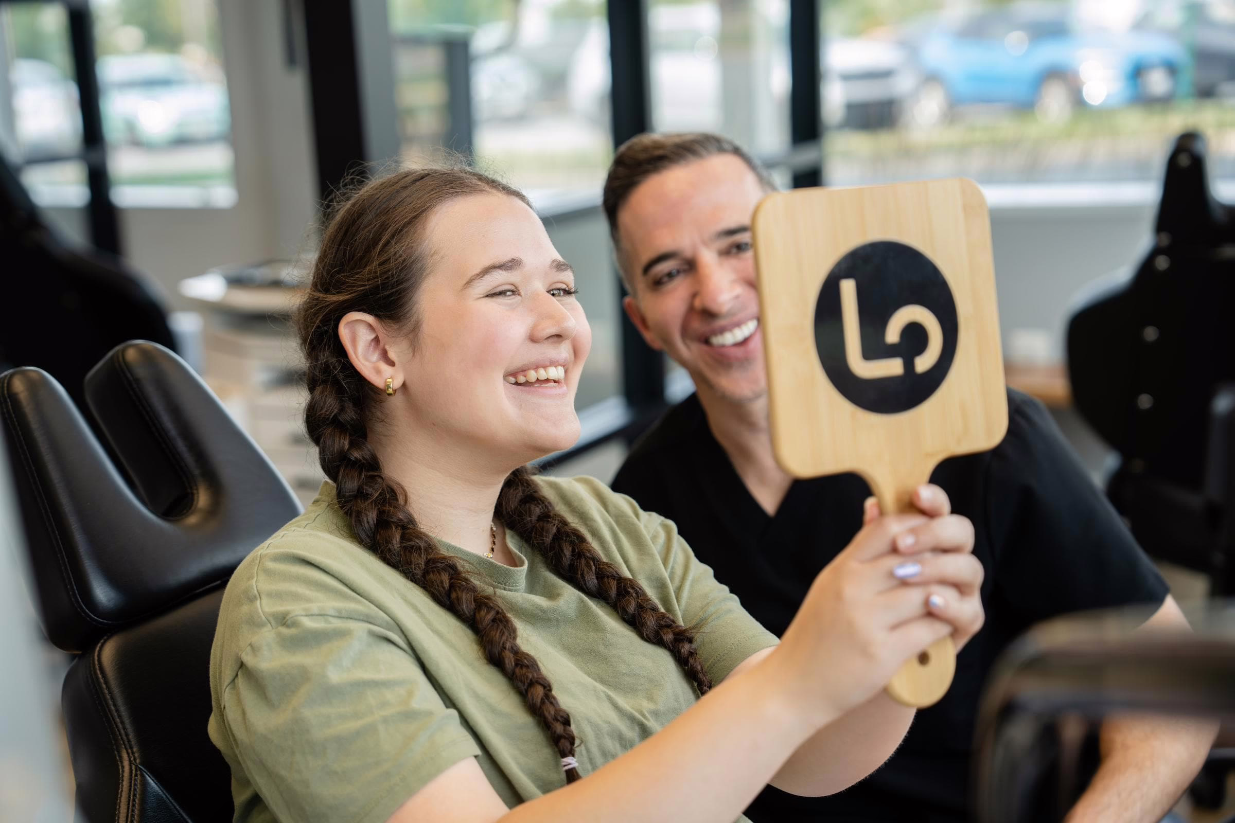 Smiling teen patient with braided hair laughs while an orthodontist holds up a wooden Legacy Orthodontics logo paddle in the Leesburg, VA office